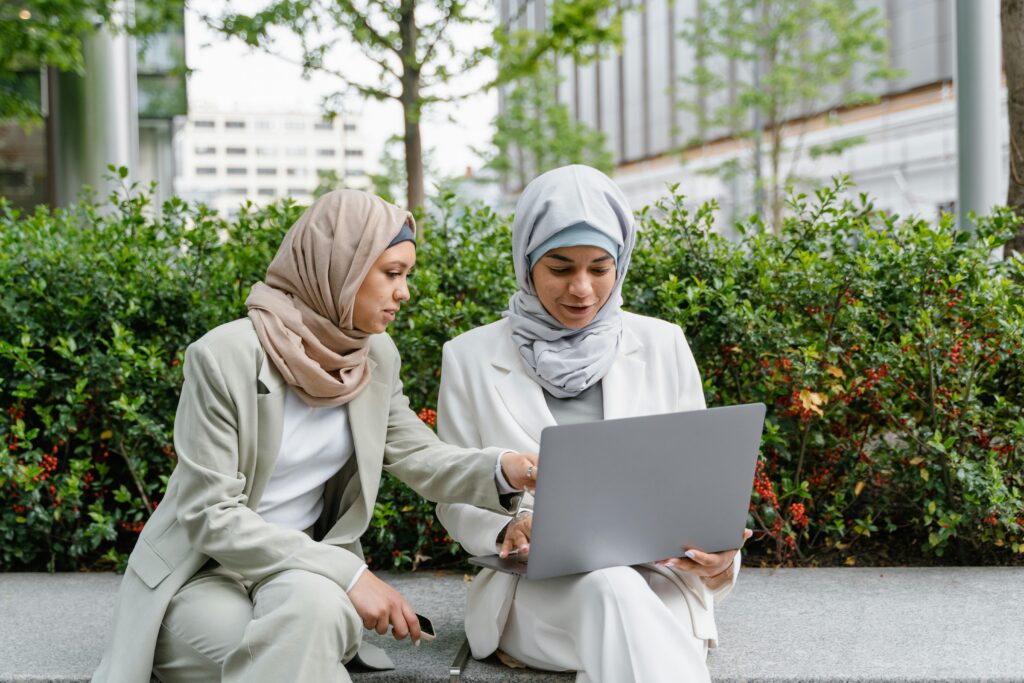 Two Muslim women in business attire working together outside on a laptop, focusing on collaboration and technology.